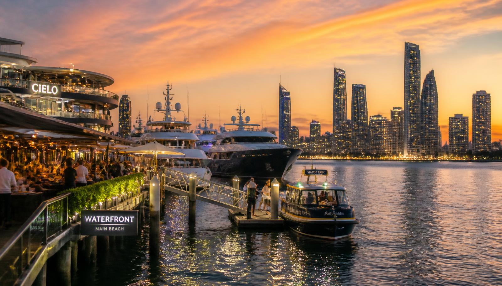 Water taxi arriving at a lit Gold Coast marina restaurant boardwalk at sunset with the Surfers Paradise skyline behind