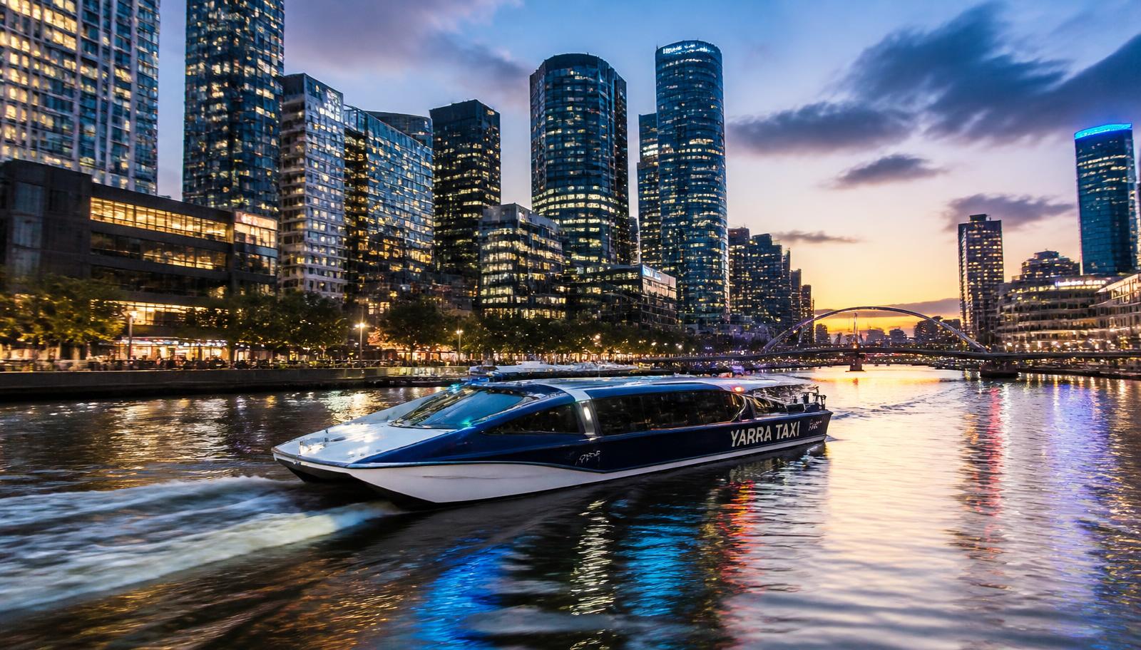Navy and white water taxi speeding along the Yarra River at dusk past the lit Southbank and Docklands towers in Melbourne